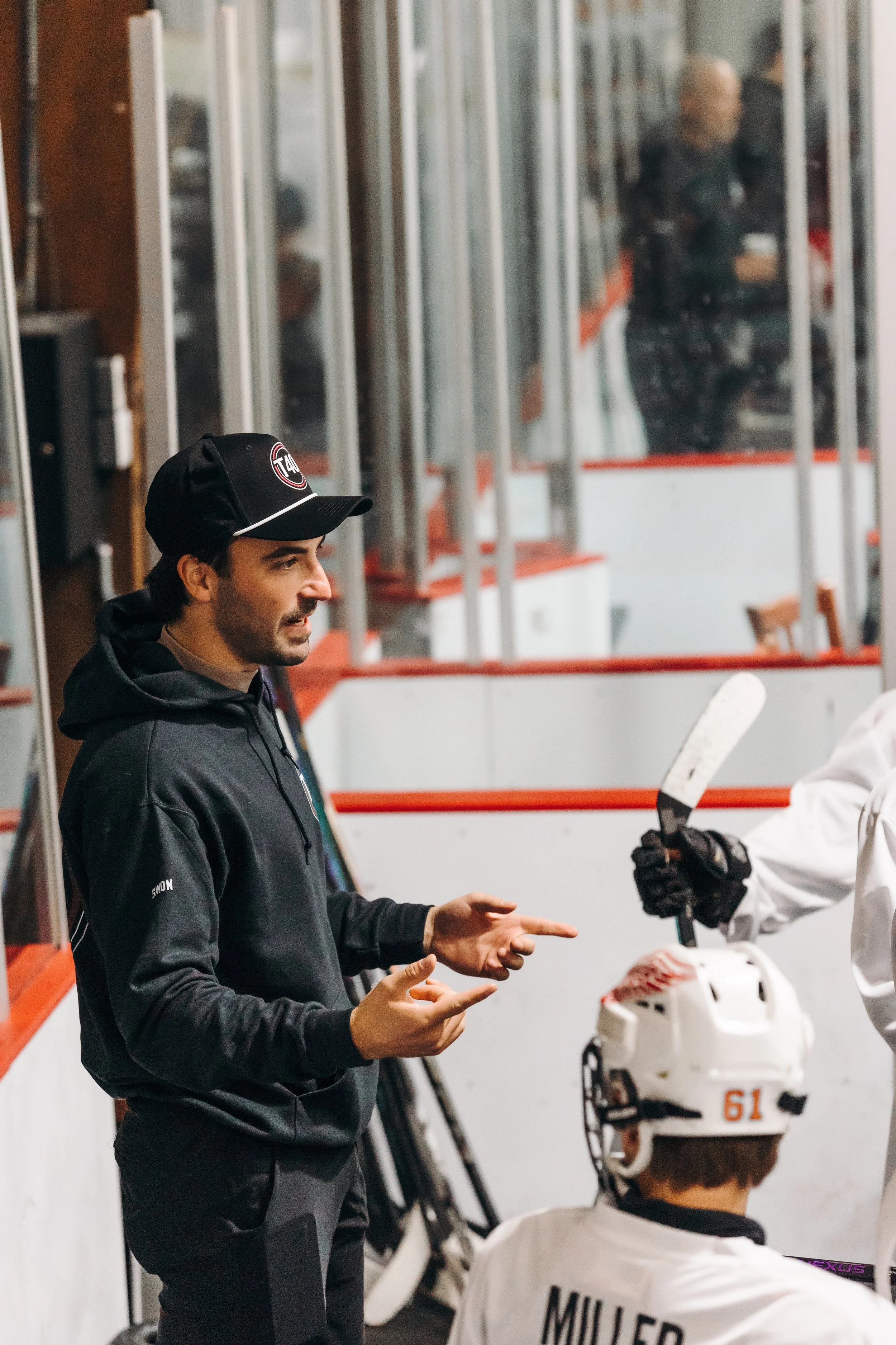 A man in a black jacket and cap standing and talking to a hockey player wearing a white jersey and helmet in a rink.