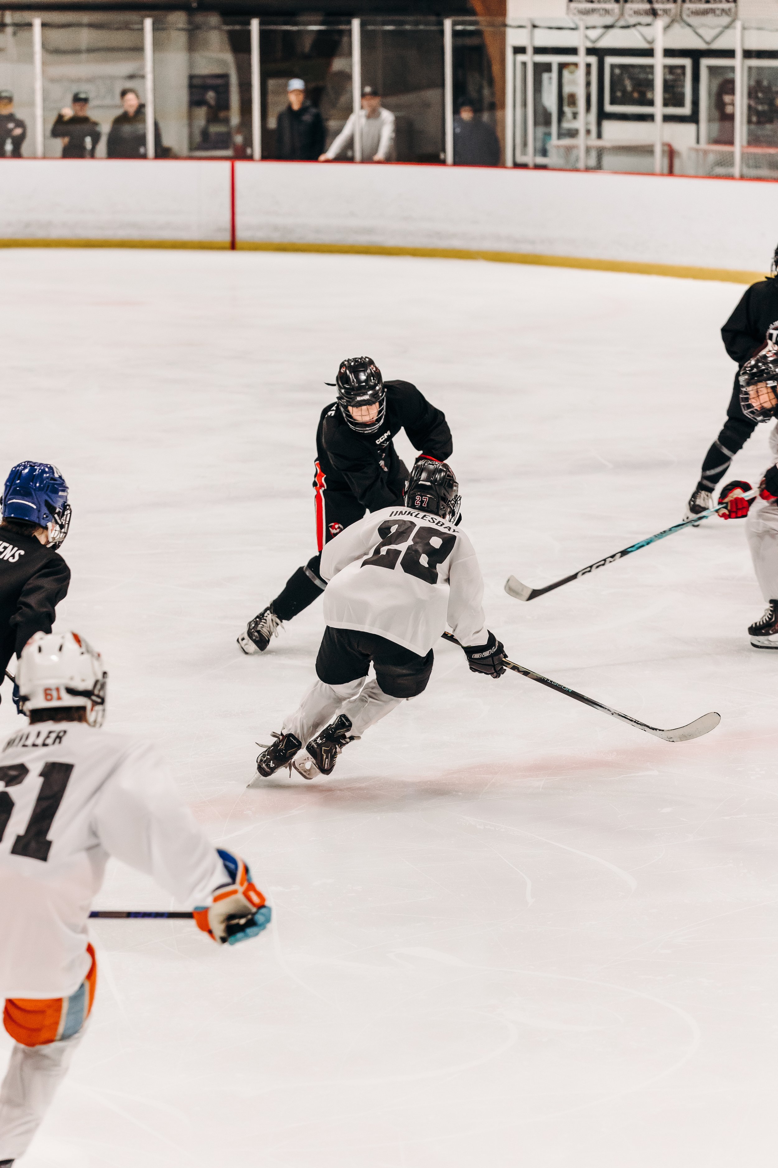 Hockey players in black and white jerseys compete on the ice rink, with players skating and tackling for the puck, while spectators watch from the bench area.