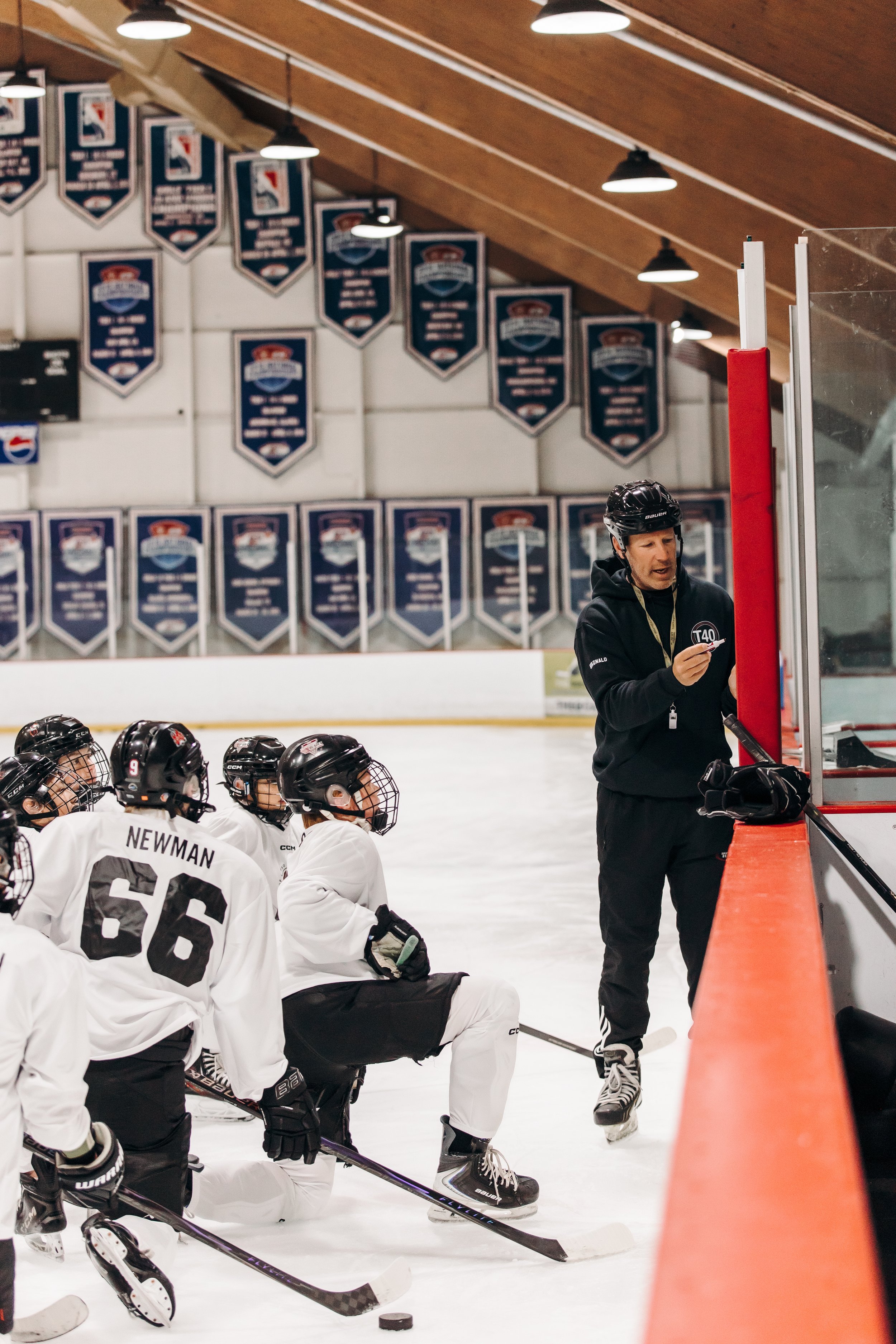 Hockey coach instructing a team of young hockey players on the ice rink during practice, with banners hanging on the wall in the background.