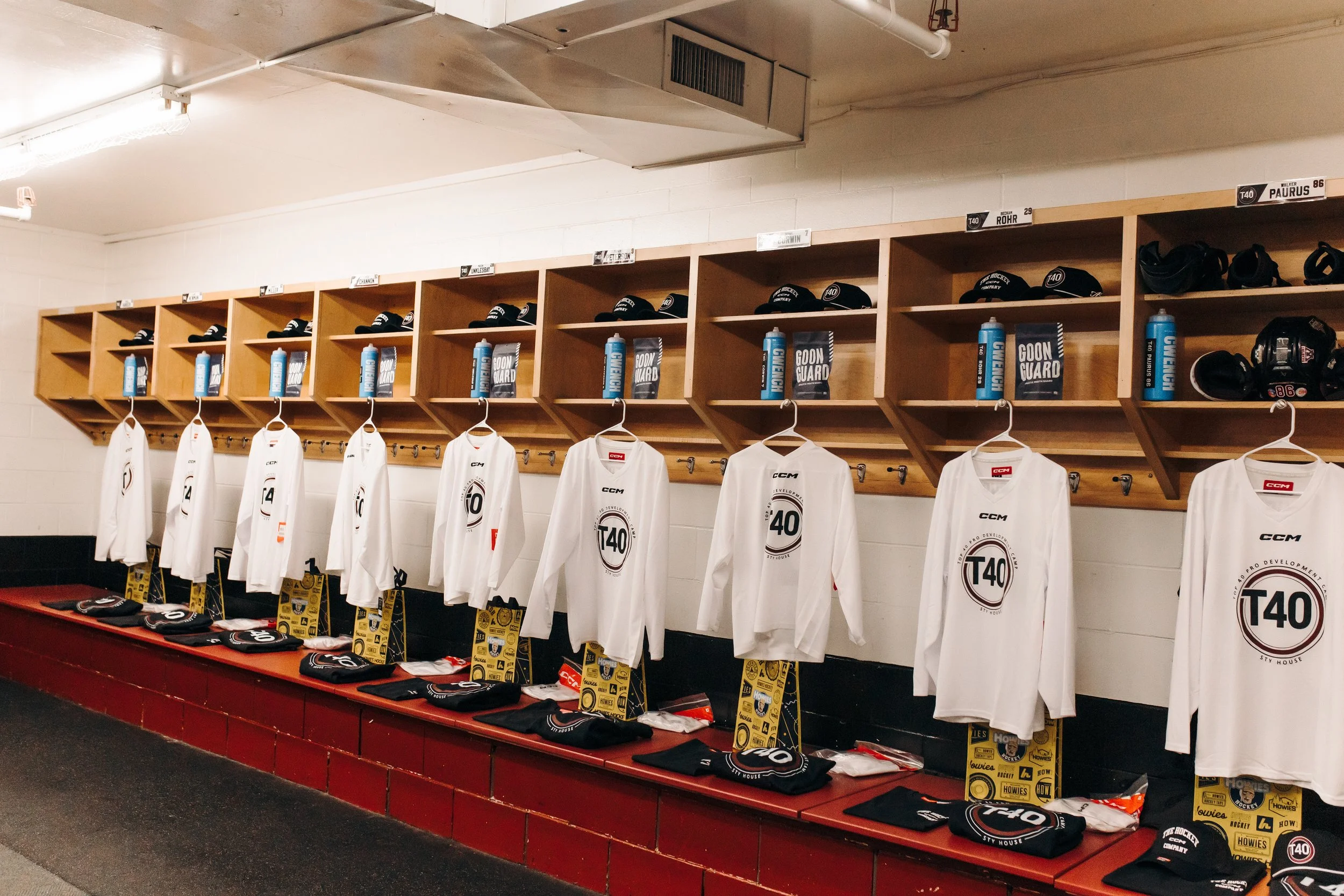 Hockey locker room with white jerseys hanging on hooks, and shelves above holding black hockey helmets, gloves, and water bottles. The jerseys display the T40 logo and player numbers, with a red bench below.