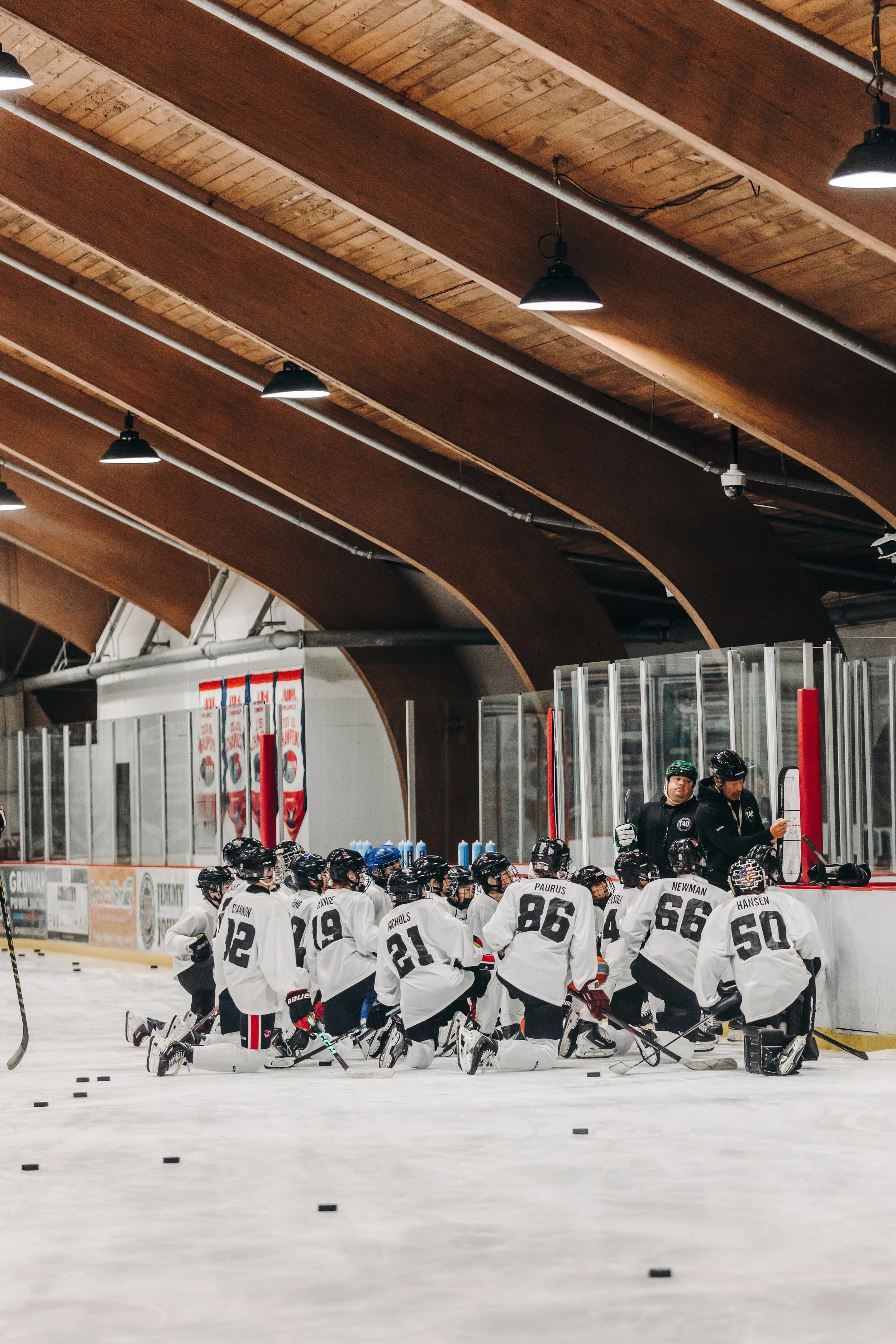 A group of hockey players kneeling on the ice rink, listening to their coach during a practice or game. The players are wearing white jerseys with black helmets and gloves. They are facing the coach, who is standing behind the glass barrier, wearing black clothing and a helmet.