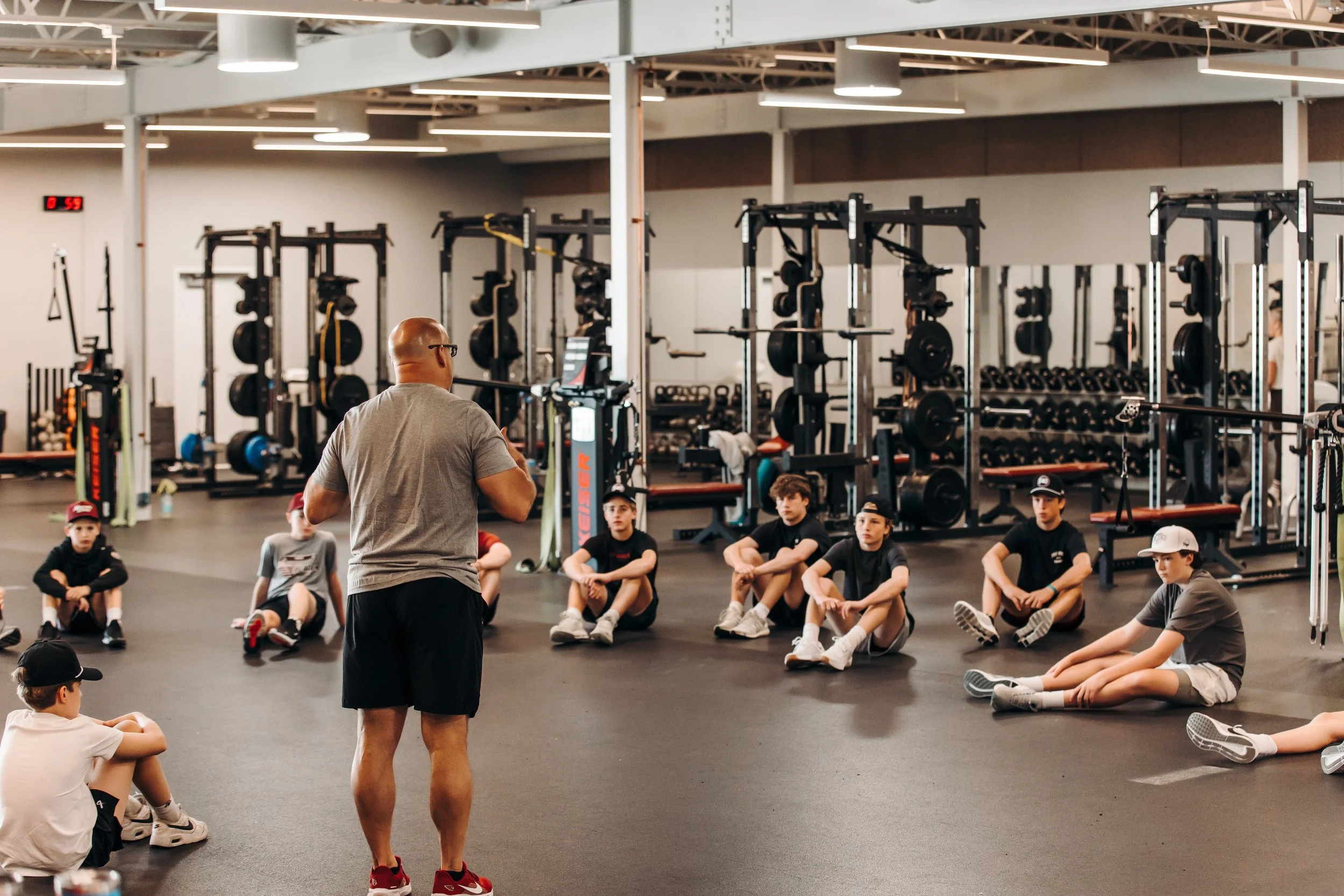 A fitness trainer is speaking to a group of children sitting on the floor of a gym. The gym is equipped with weightlifting equipment and has a digital clock on the wall.