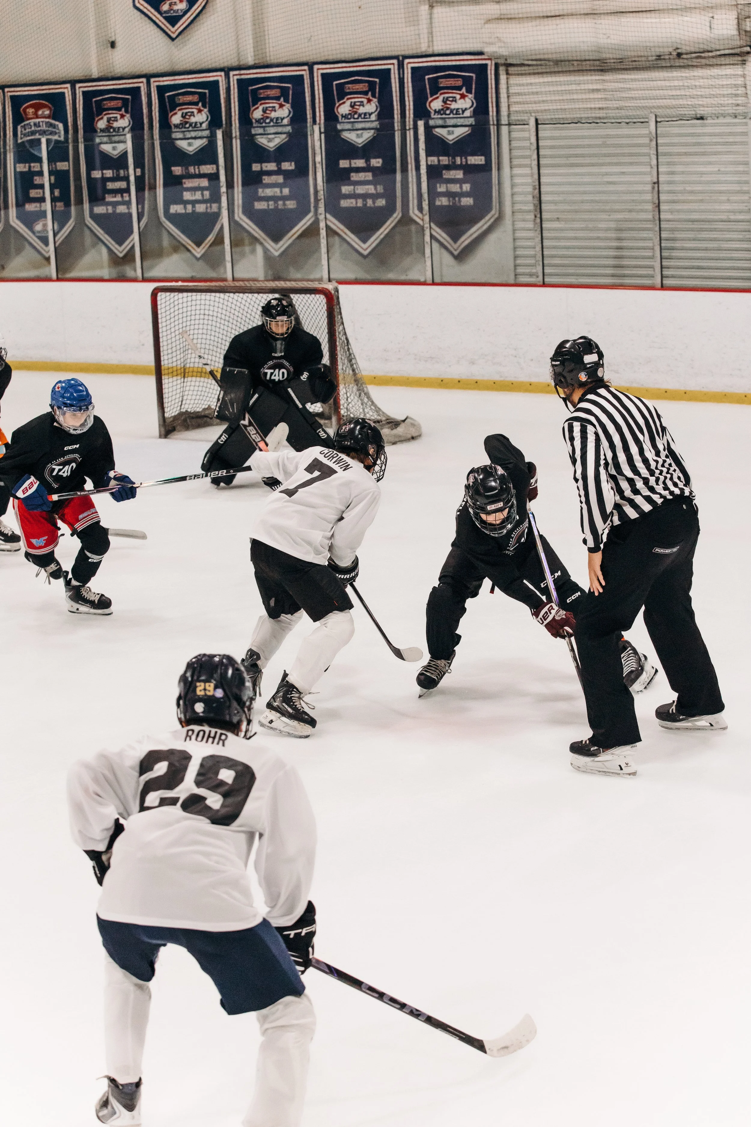 Ice hockey players in black and white jerseys compete for puck, with referee observing, in an ice rink with banners hanging on the wall.