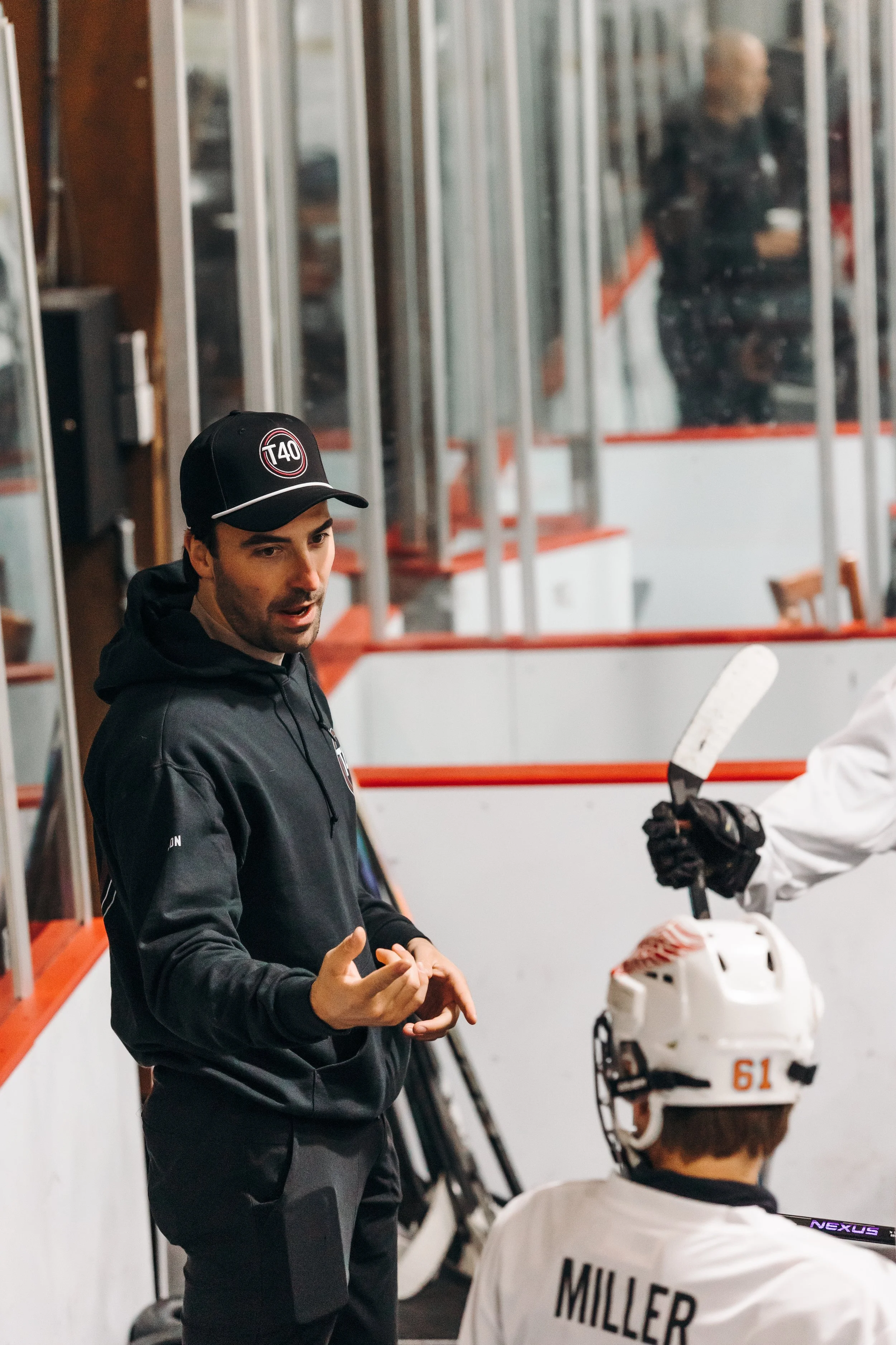 A man wearing a black baseball cap with a red and white logo and a black jacket, talking to a person in hockey gear holding a hockey stick. The setting appears to be an indoor ice rink with glass panels and benches in the background.