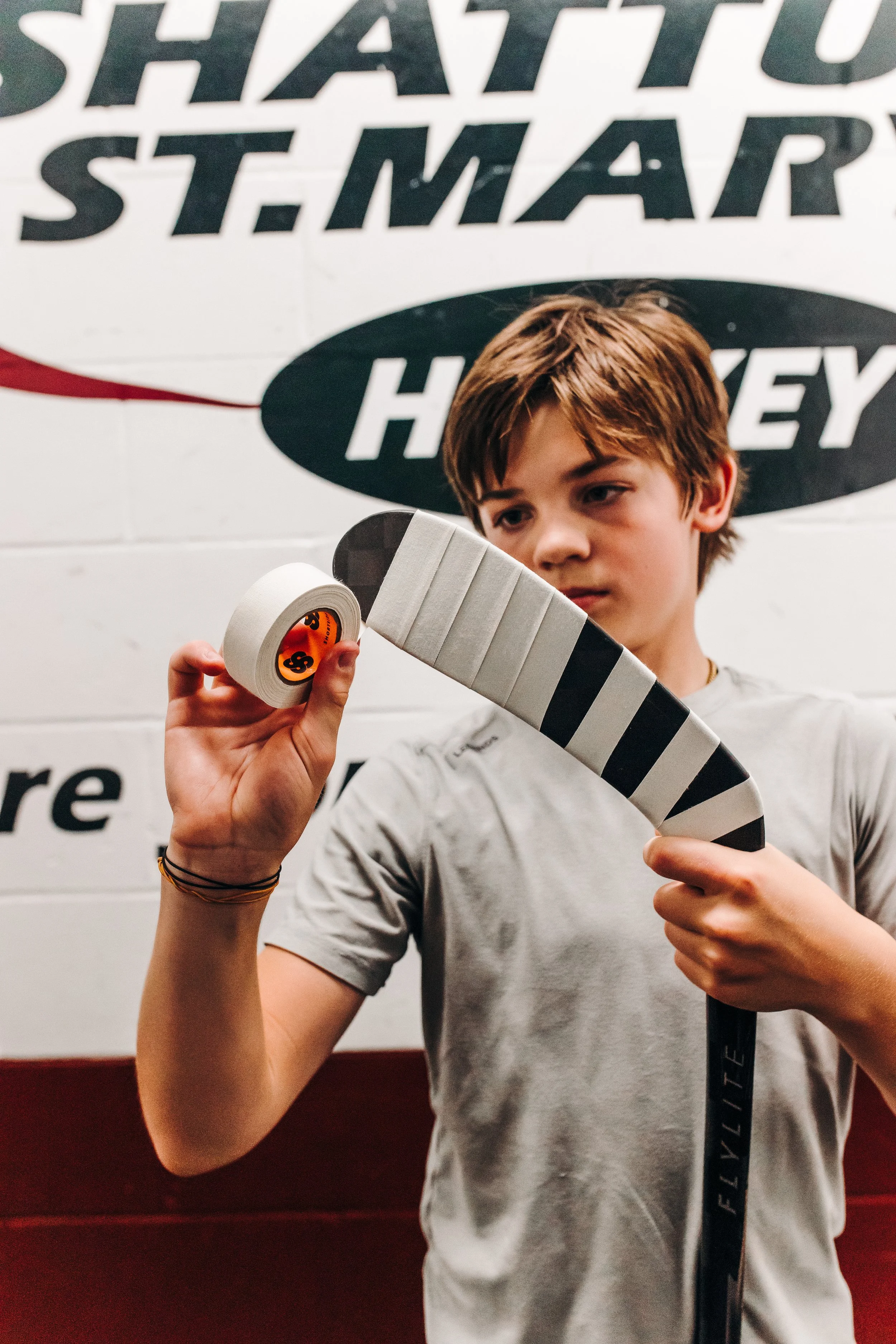 A young person with brown hair holding hockey tape and a hockey stick, with a hockey-themed sign in the background.
