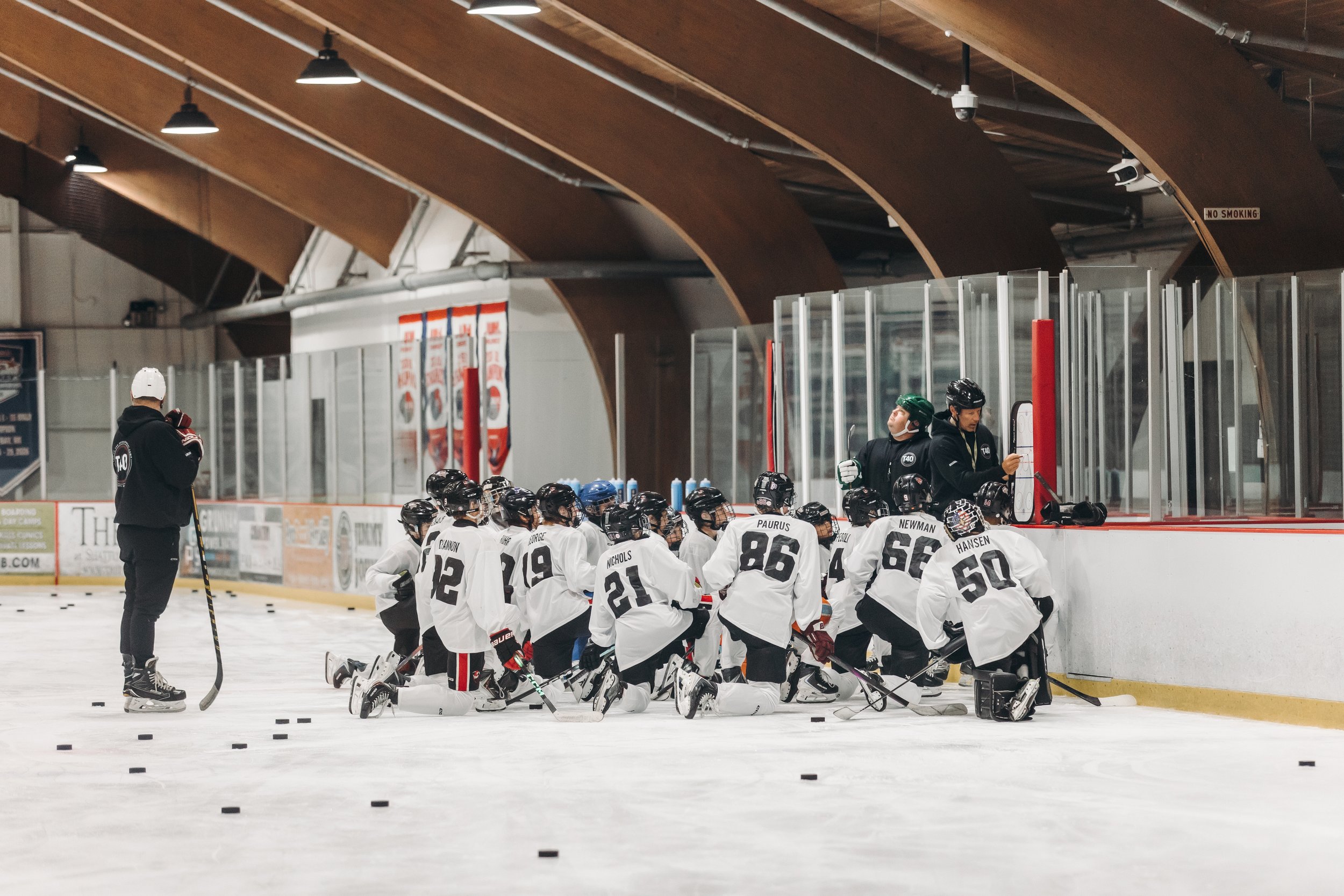 A group of young ice hockey players kneeling on the ice rink, listening to their coaches during practice or a team meeting. The players are dressed in white jerseys with black helmets, and the coaches are standing near the team.