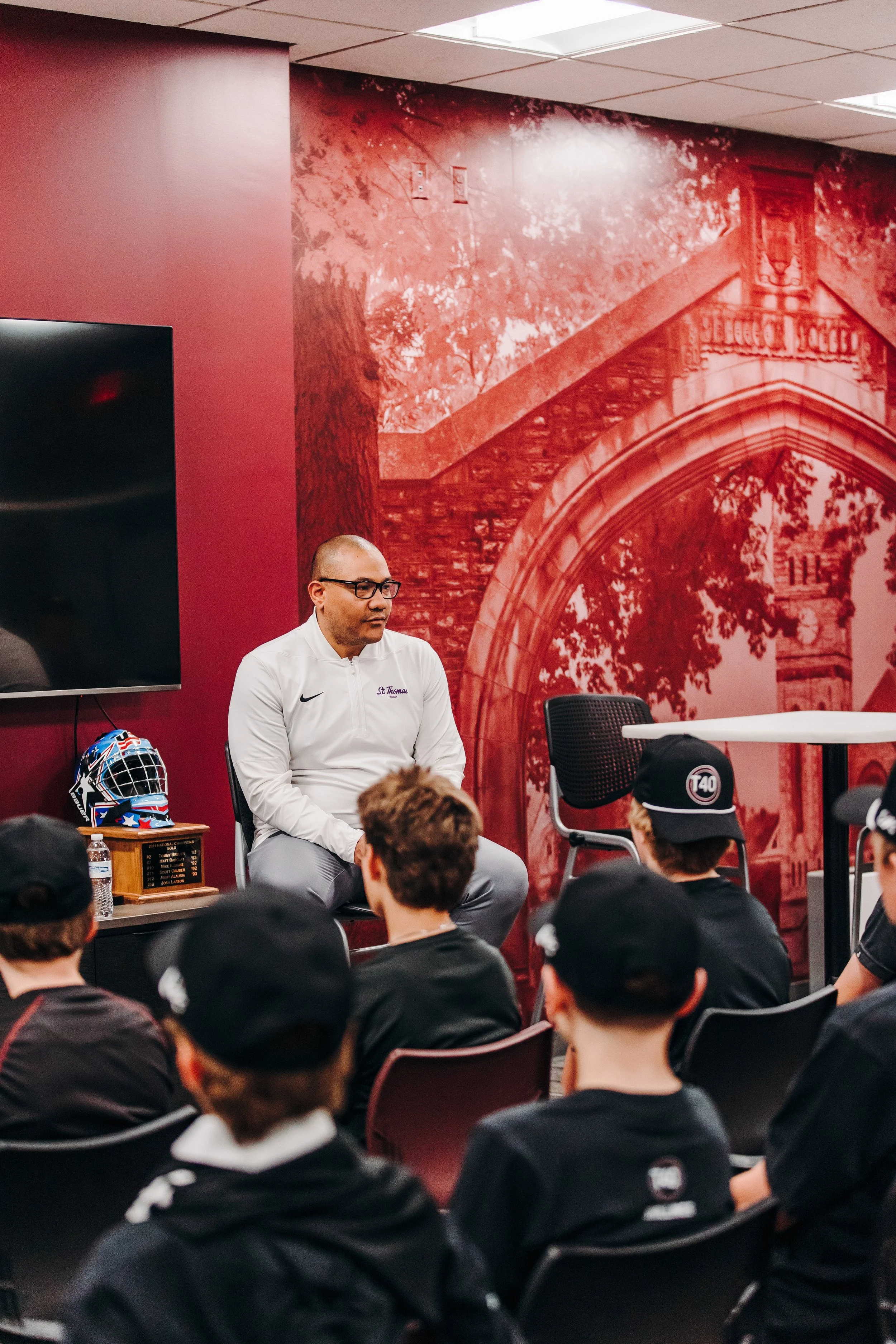 A group of young ice hockey players with black jerseys and caps watching a coach during an indoor team meeting. The coach, wearing a white shirt with glasses, is seated in front of them. The background features a large red mural with trees and a ston
