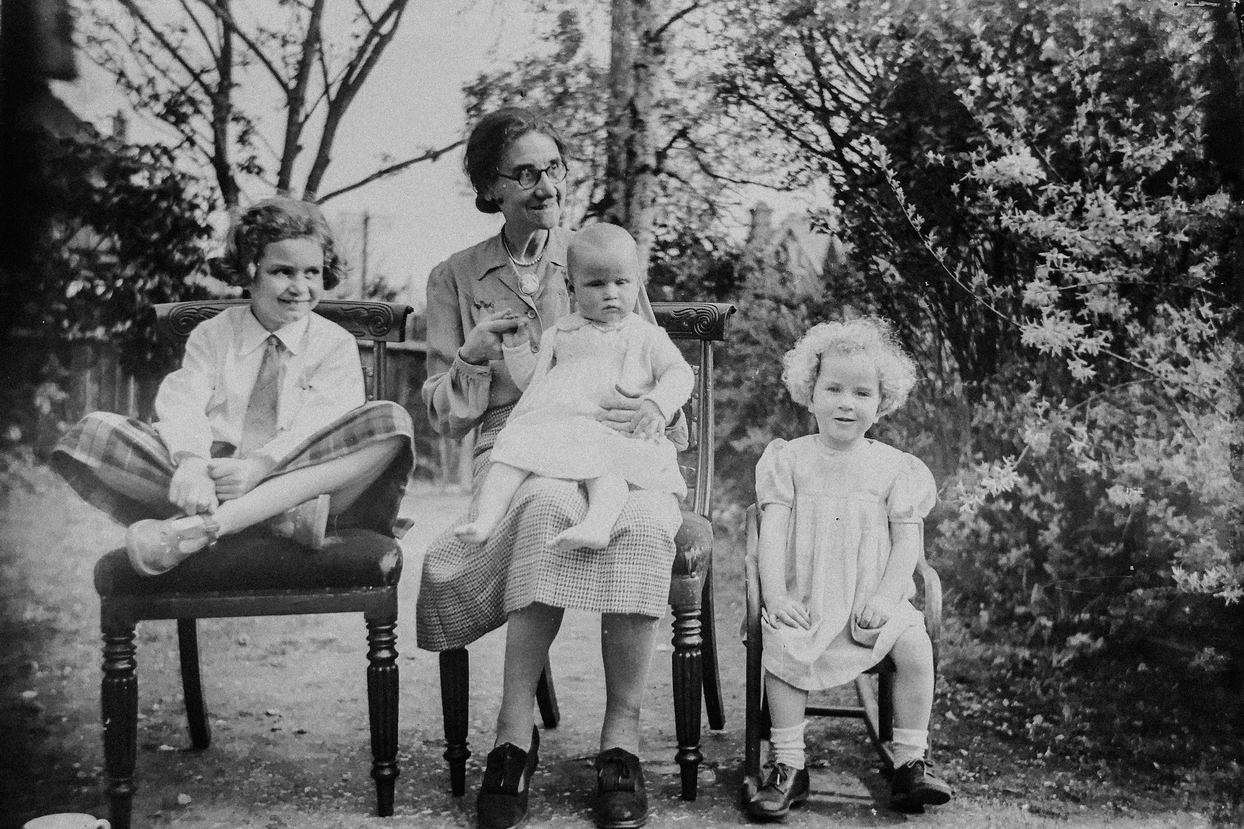 Black and white photo of four children and one adult woman outdoors. Two children stand near a small table, one woman and one young girl sit on chairs, and trees and foliage are in the background.