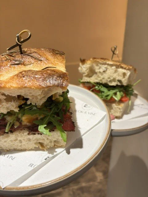 Close-up of a sandwich with arugula and other greens, served on a white plate with a line of parchment paper underneath, and another similar sandwich in the background on a separate plate.