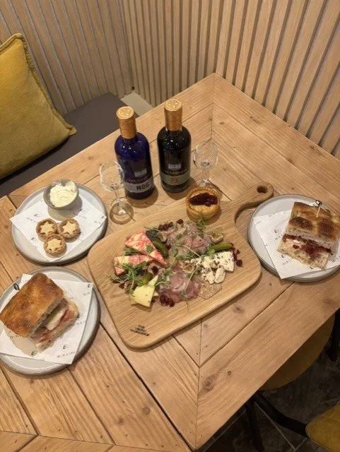 A wooden table with two plates of sandwiches, a wooden serving board with sliced cured meat, cheese, and greens, two bottles of wine, and small glasses, along with a bowl of butter and some biscuits on a white plate.