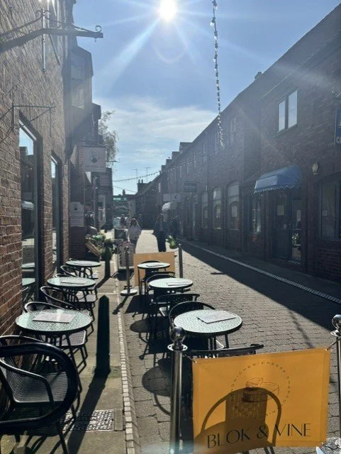 Outdoor seating with black metal chairs and round tables on a cobblestone sidewalk. A yellow sign for "Blok & Vine" in front, and buildings lining the street with shops and windows on a sunny day.