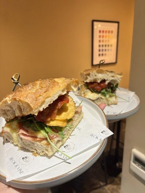 Two sandwiches on white plates with various fillings, held together with toothpicks, in a cozy indoor setting.