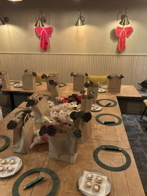 A long wooden table set with paper gift bags, each with a black honeycomb bow and some containing wrapped gifts or crafts. The table has green paper napkins and some plates with small pastries. The background features a wall with three pink bow decor