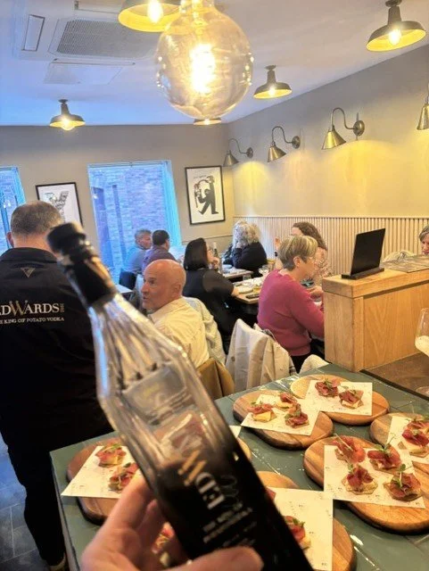 Inside a busy restaurant with customers sitting at tables, some watching a computer, with modern lighting and framed artwork on the wall. In the foreground, a hand holds a bottle of beer, and wooden serving boards with appetizers are on the table.