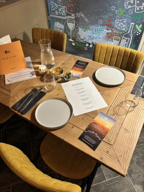 A dining table set for four with white plates, wine glasses, water glasses, black napkins, and a menu or program, decorated for a celebration with Christmas-themed wall art in the background.