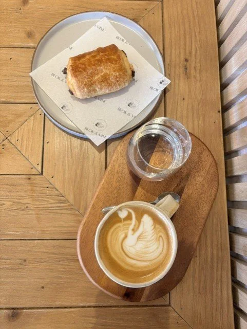 A wooden table with a round metal plate holding a grilled sandwich with toasted bread, a glass of water, and a latte art coffee on a wooden serving board.