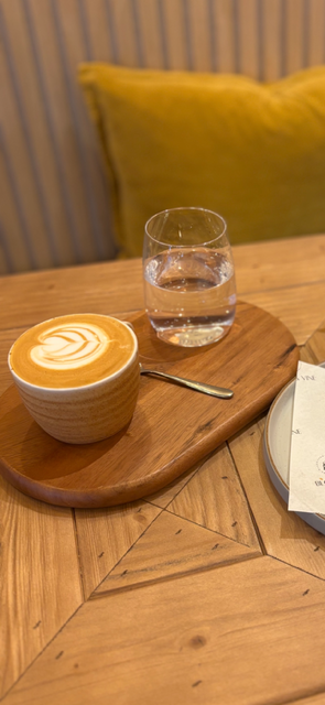 A cup of coffee with foam art, a glass of water, and a spoon on a wooden serving tray on a table with a yellow pillow in the background.