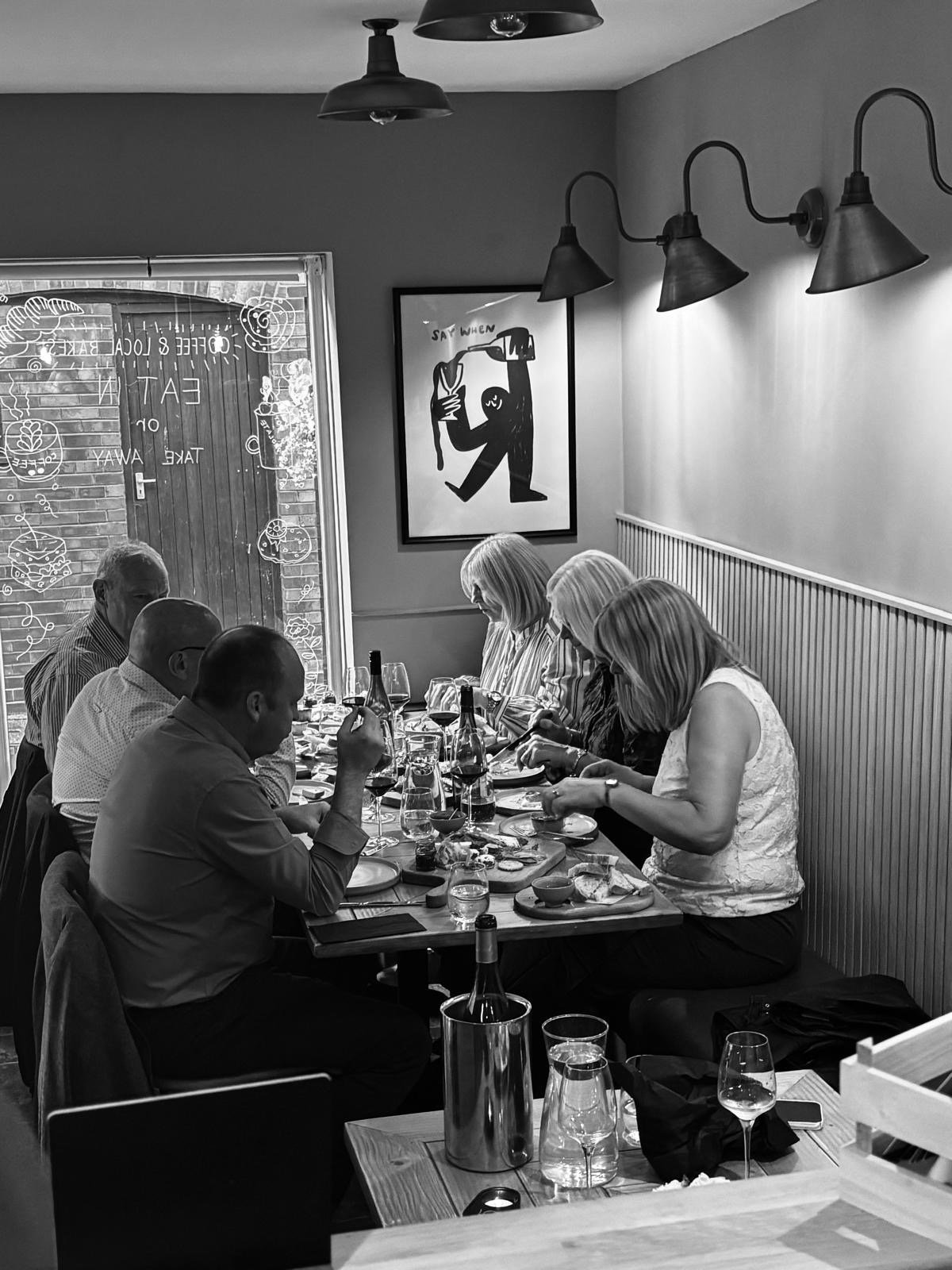 A group of six people sitting at a dining table inside a restaurant, with plates, glasses, and bottles of wine. The restaurant has a gray interior with a window, wall art, and wall-mounted lamps.