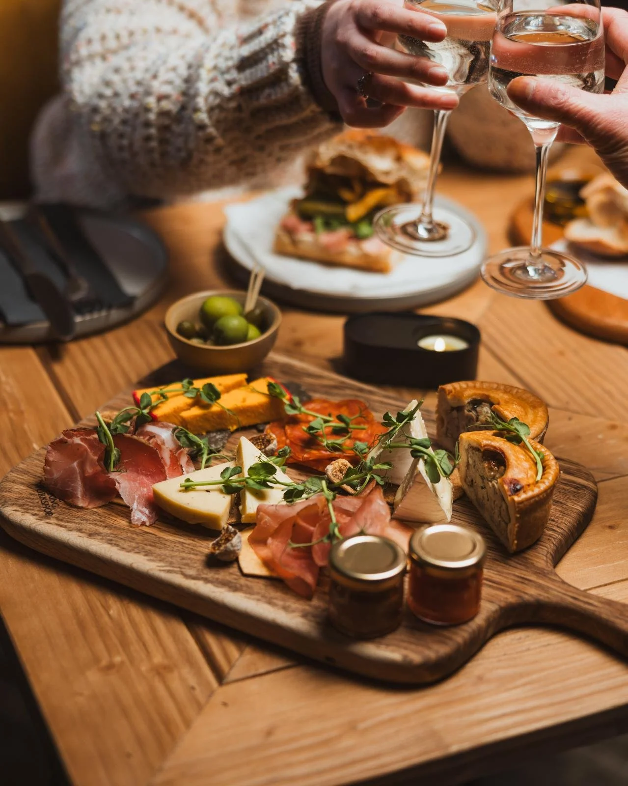 A charcuterie platter with assorted cheeses, cured meats, and garnishes on a wooden board, surrounded by small jars, green olives, and other appetizers. People are clinking glasses of wine in the background.