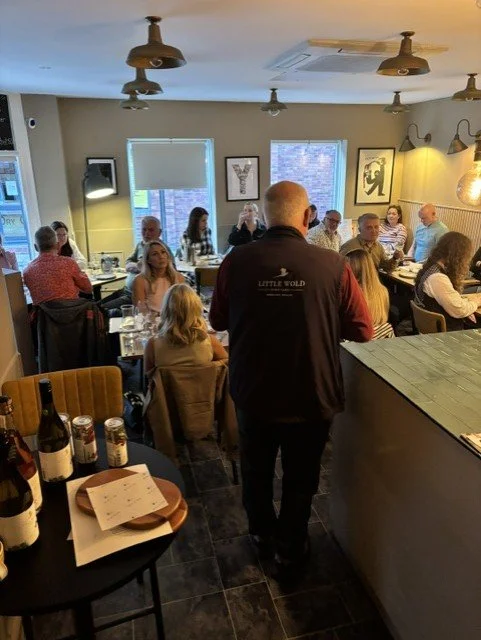 People dining in a restaurant with several tables filled with guests and a man standing in the foreground facing the group.