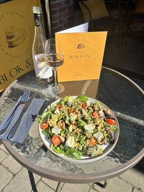 A round glass table with a fresh salad, a glass of white wine, a bottle of sparkling water, a blue napkin with a fork and knife, and a yellow menu from Blok & Vine, outdoors with chairs and brick wall in background.