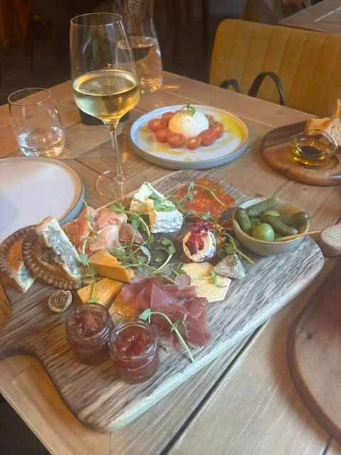 A wooden serving board with assorted cheeses, cured meats, olives, and small jars of jam or preserves. In the background, there is a glass of white wine, a small bowl of tomato and mozzarella salad, and additional glasses and dishes on a wooden table