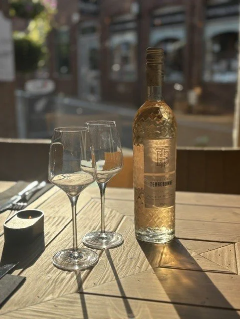 A bottle of white wine and two empty wine glasses on a wooden table outside, with sunlight casting shadows, and urban buildings in the background.