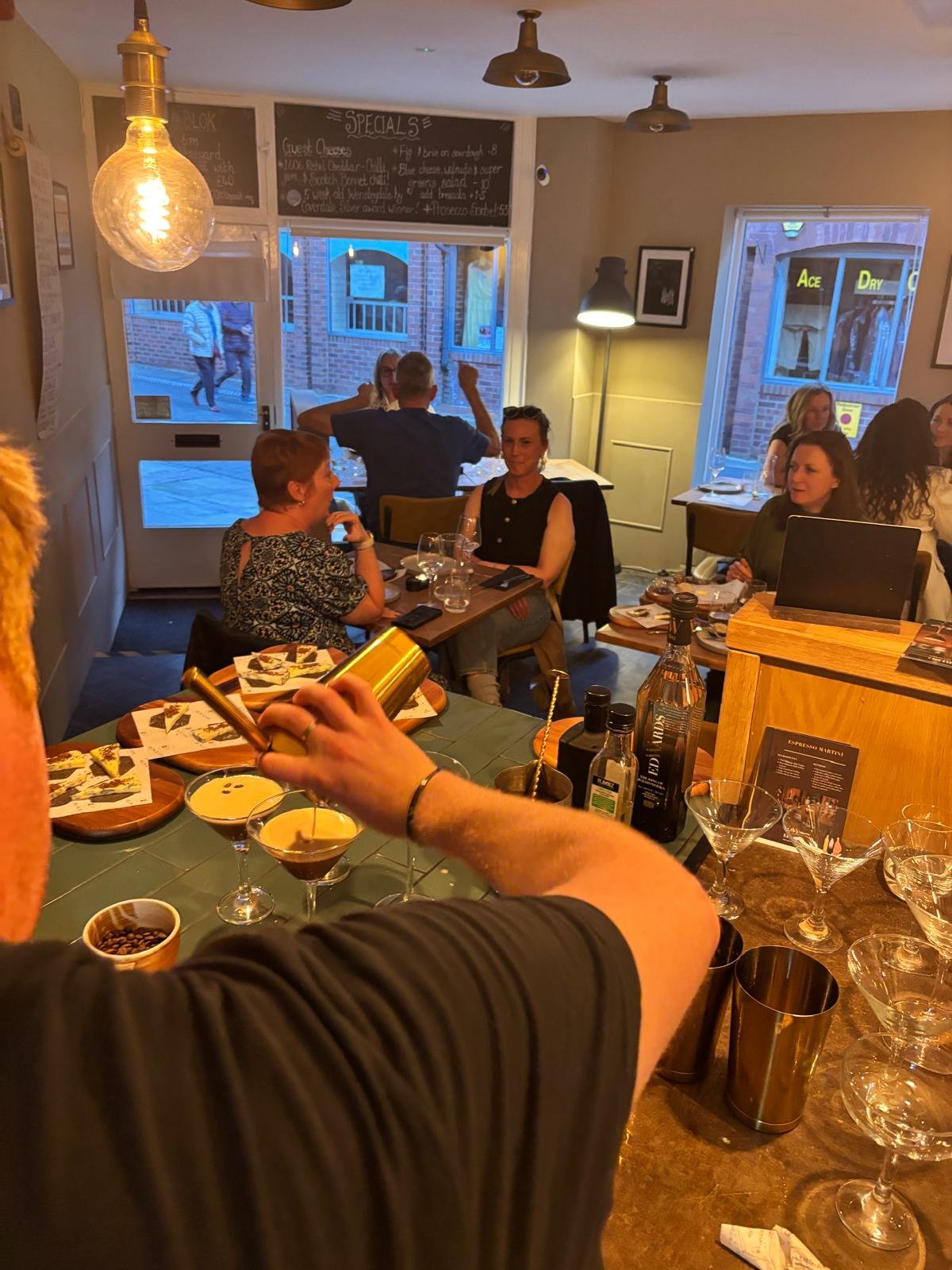 People sitting at tables in a cozy restaurant, with a bartender pouring drinks, various bottles and glasses on the bar, and a chalkboard menu above the door.