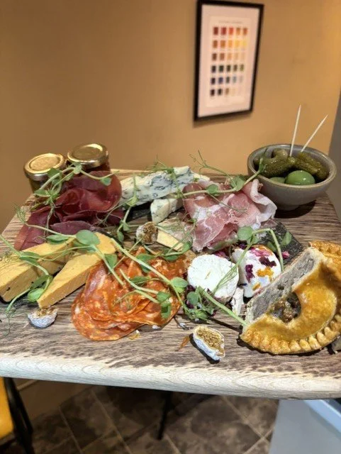 A charcuterie board with cheese, cured meats, olives, cheese dips, and a small bowl of pickles.