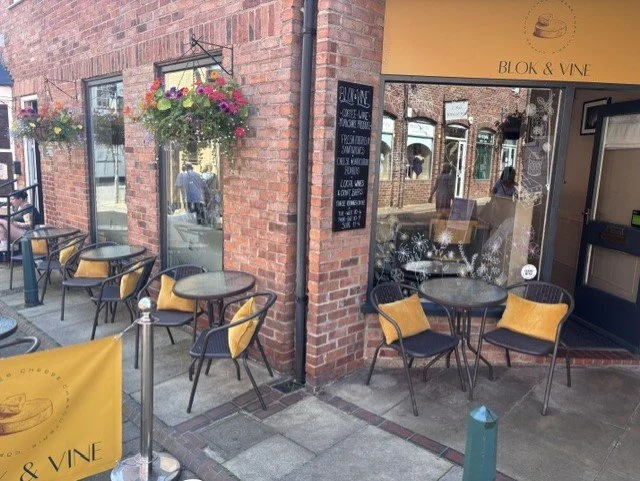 Outdoor seating area of a cafe with yellow chairs and black tables outside a brick building with hanging flower baskets and a blackboard menu, part of Blok & Vine restaurant.