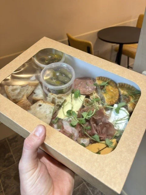 Hand holding a pizza box with a clear plastic window, revealing assorted various toppings and sides inside, in a restaurant setting.
