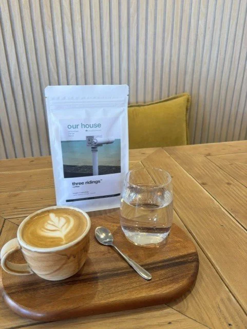 A cup of coffee with latte art, a glass of water, a spoon, and a bag of coffee beans on a wooden tray, set on a wooden table with a yellow cushion in the background.