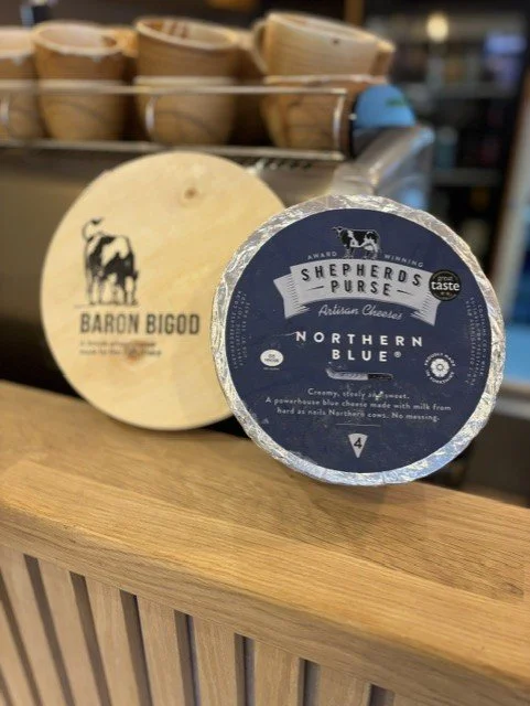 A round block of Baron Bigod cheese and a Shepherds Purse Northern Blue cheese on a wooden surface, with a background of stacked wooden baskets.