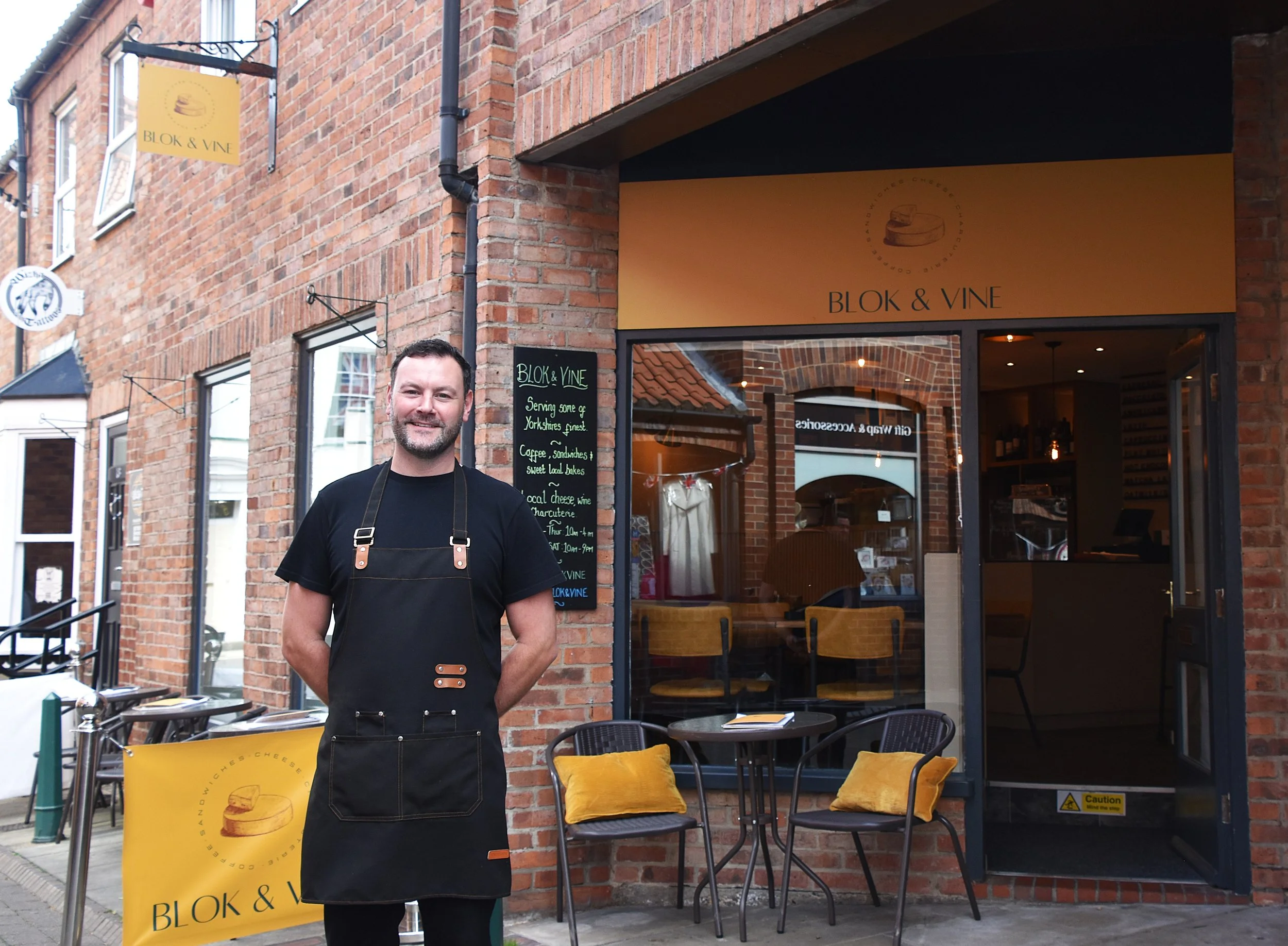 A man wearing a black apron standing outside a brick building with a sign that reads "BLOK & VINE." There are yellow cushions on black chairs and a yellow banner with the restaurant's logo. The restaurant appears to be a cafe or diner with indoor seating visible through the window.