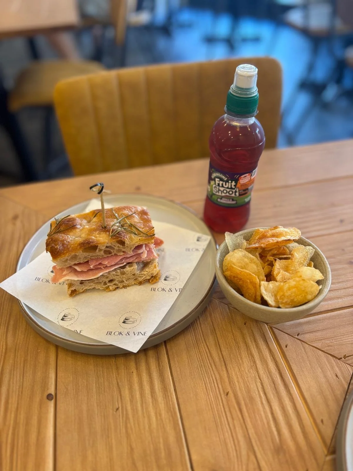 A sandwich with ham and cheese on a toasted baguette with herbs, served on a plate with paper liner from Blok & Vine. Beside it is a bowl of potato chips and a bottle of red fruit juice, all on a wooden table in a restaurant setting.