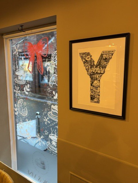 Framed artwork of the letter 'Y' with various illustrations and the word 'Yokohama' inside it, hanging on a wall next to a window decorated with a red butterfly ornament and other stickers.