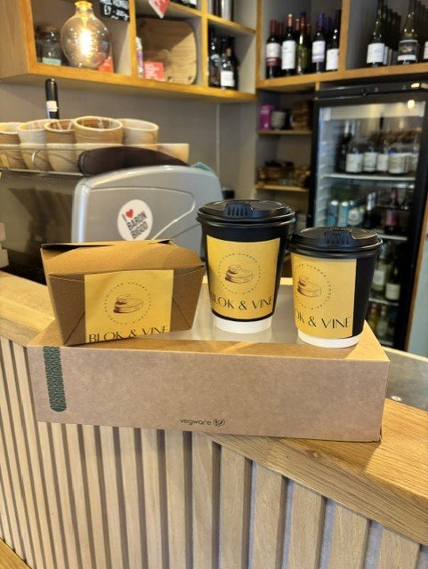 Two to-go coffee cups and a small paper bag with yellow labels from Blok & Vine on a wooden counter in a coffee shop. Shelves filled with wine bottles and a refrigerator are in the background.