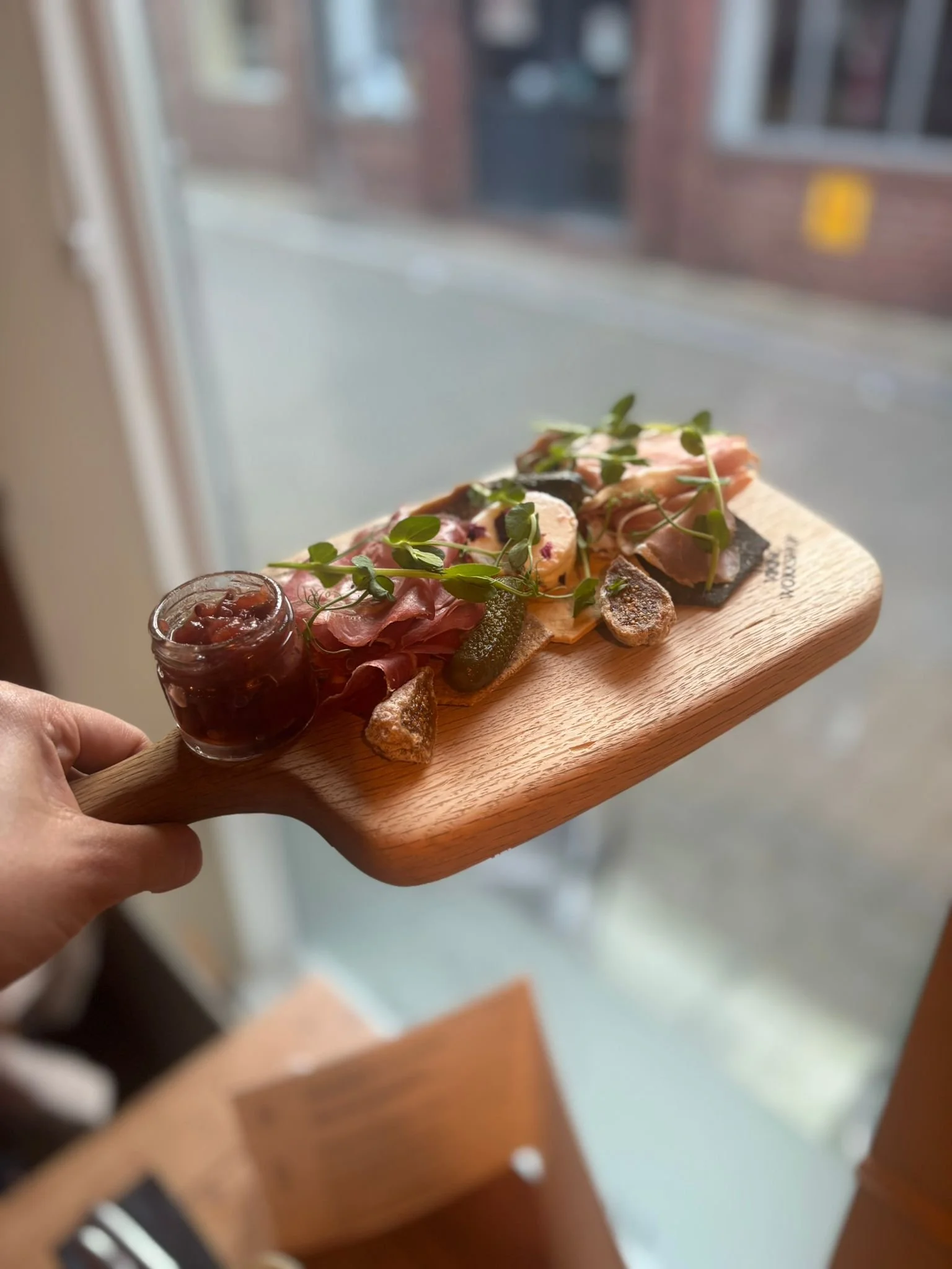 A person holding a wooden serving board with assorted cured meats, microgreens, and a small jar of jam or preserves near a window.