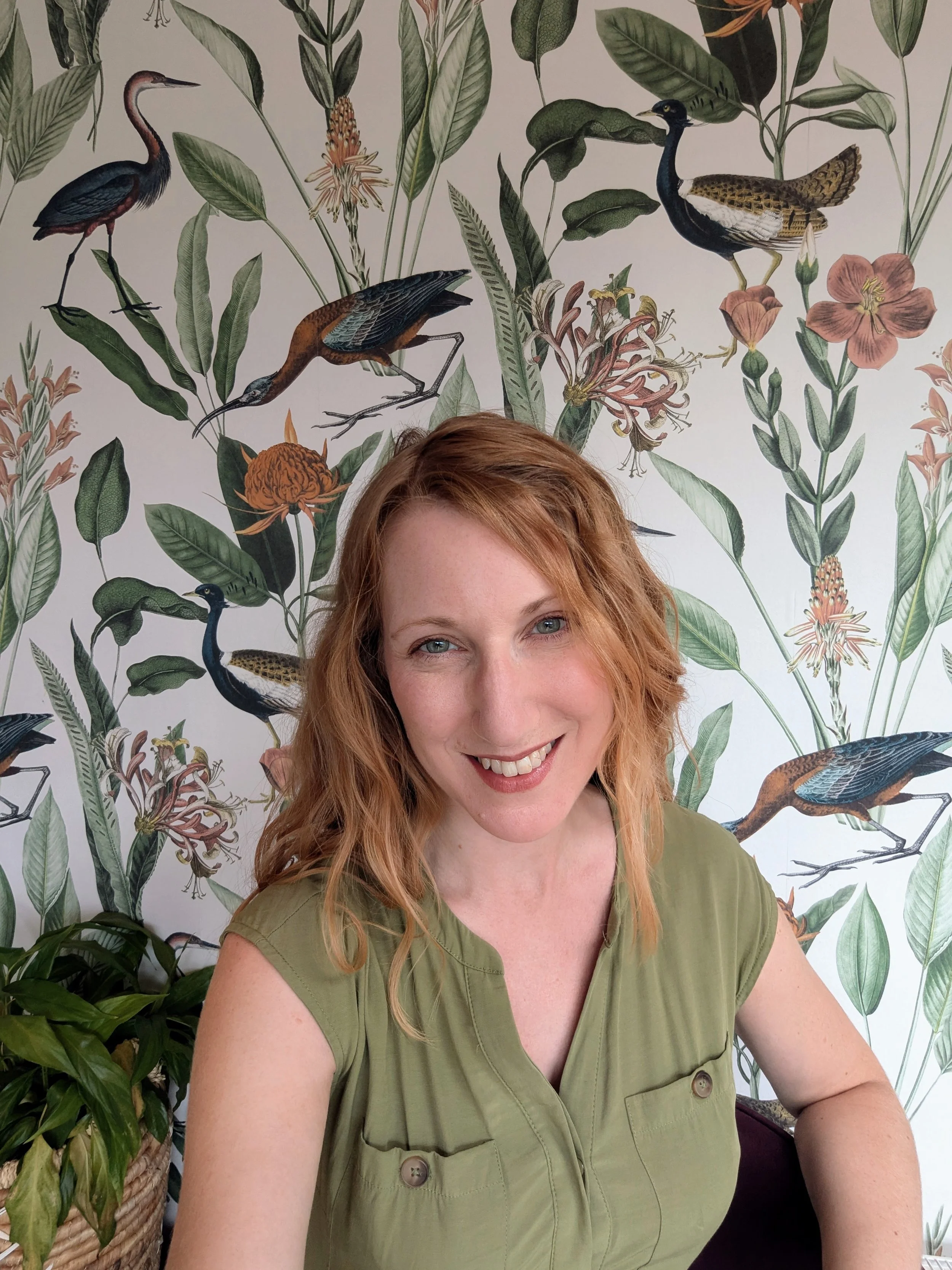 A woman with red hair and a green shirt smiling in front of a wallpaper with illustrations of flowers and various birds.