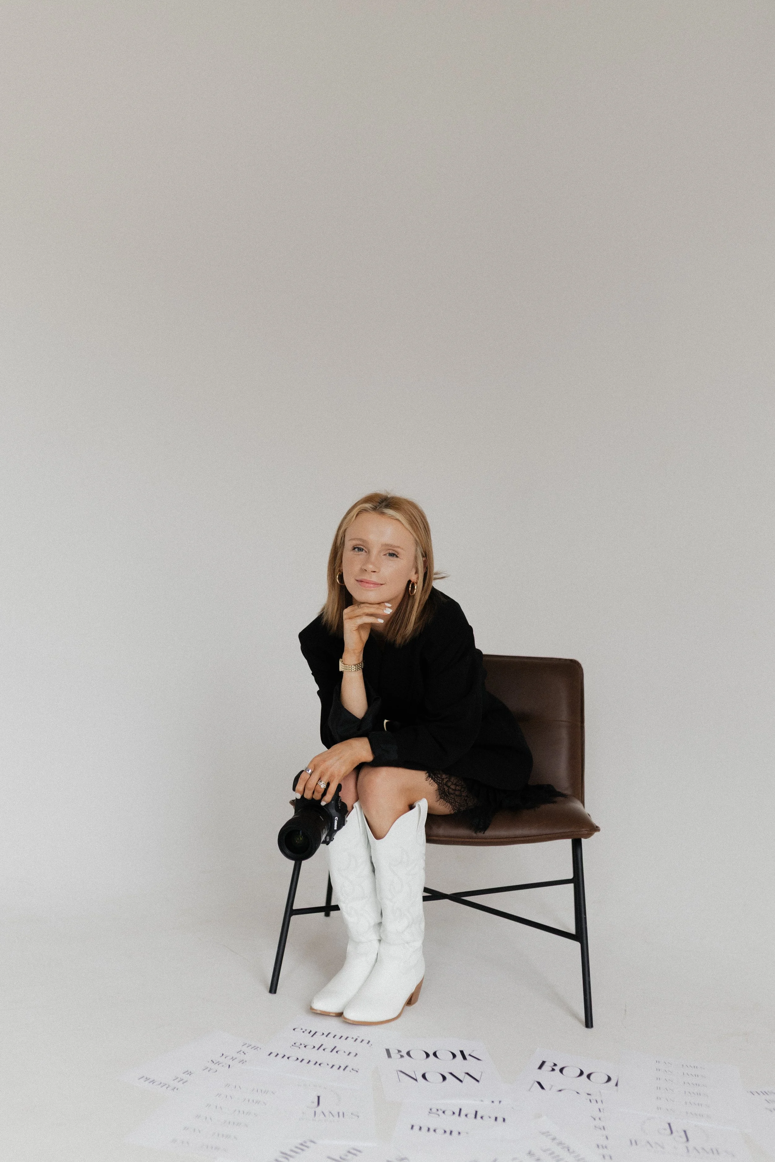 A woman seated on a chair with a camera, wearing white boots and a dark outfit, in front of a plain background with scattered text papers on the floor.