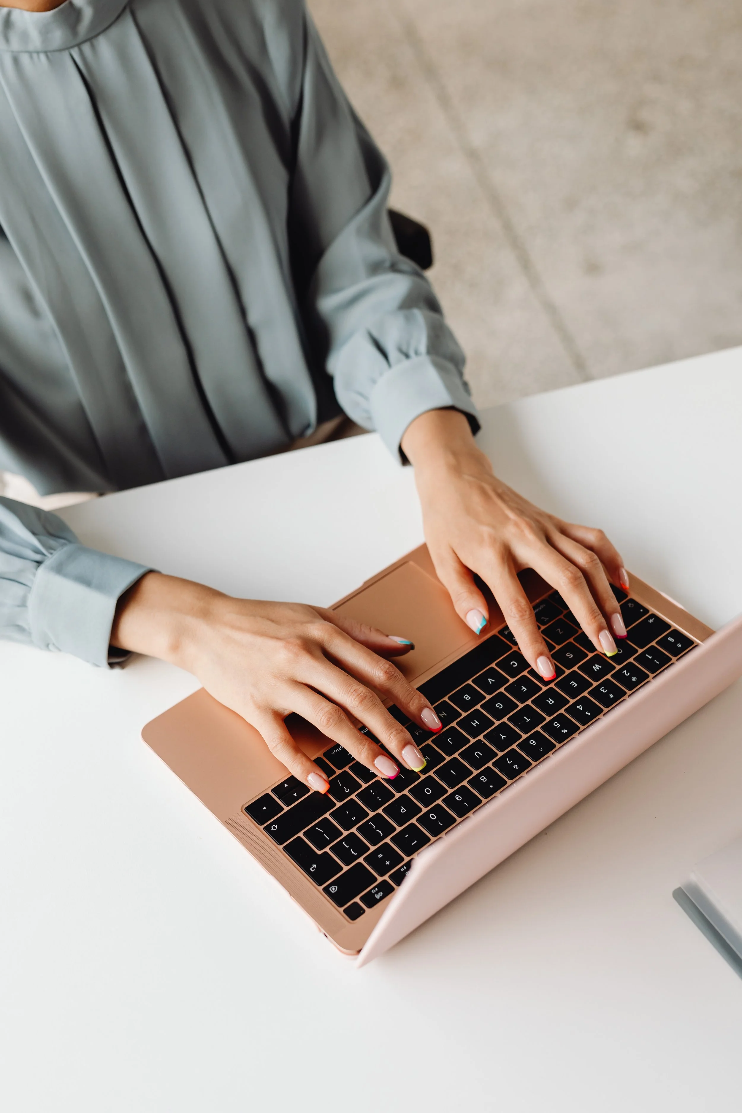Person typing on a pink laptop with colorful nails at a white table.