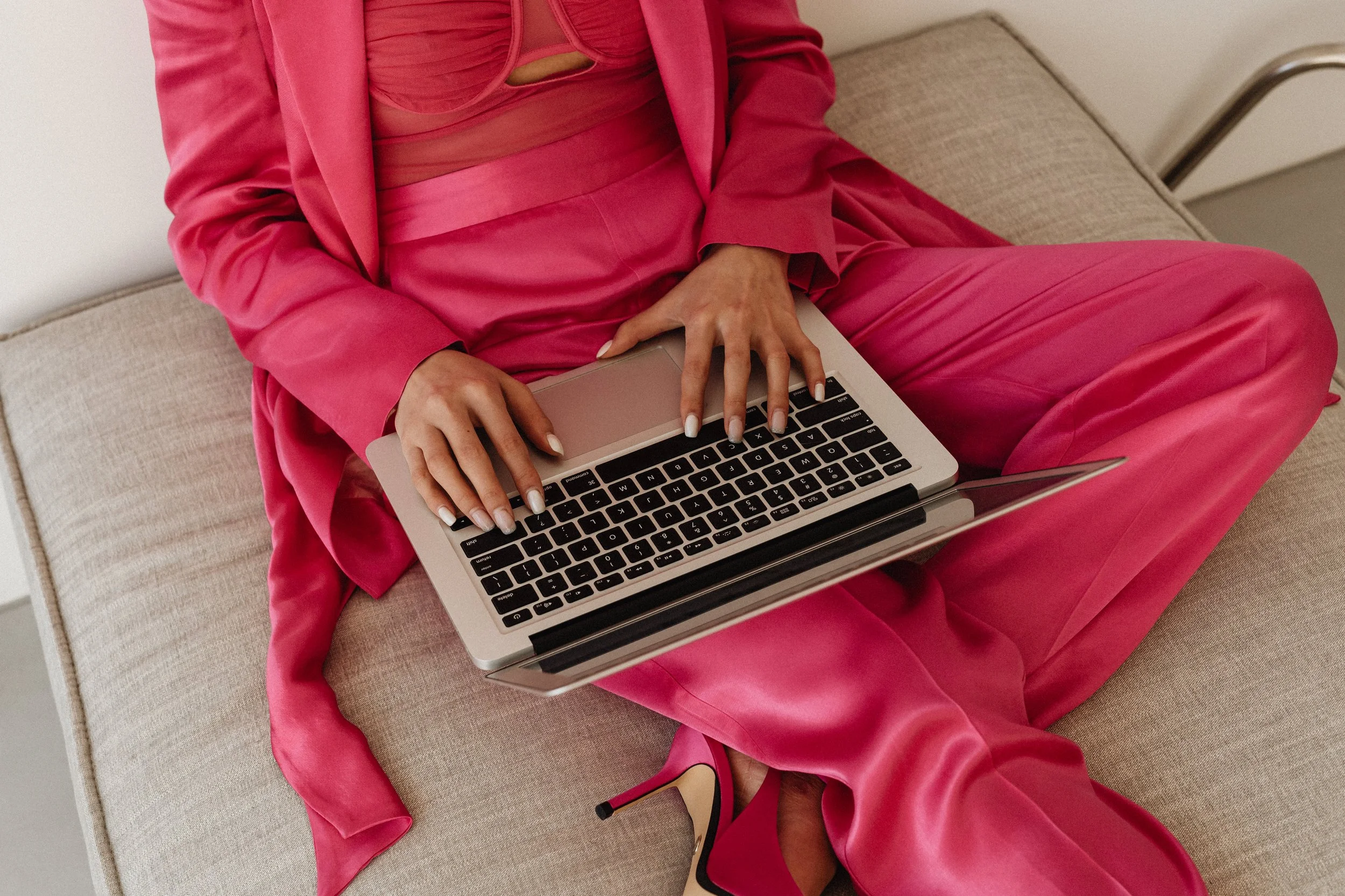 A person in pink clothing sitting on a beige couch using a laptop.