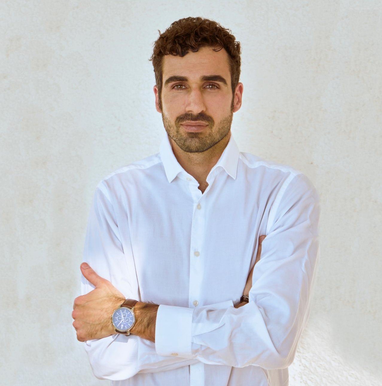 Hombre con barba y cabello rizado, vestido con camisa blanca, cruzado de brazos, usando reloj en la muñeca izquierda, frente a fondo claro.