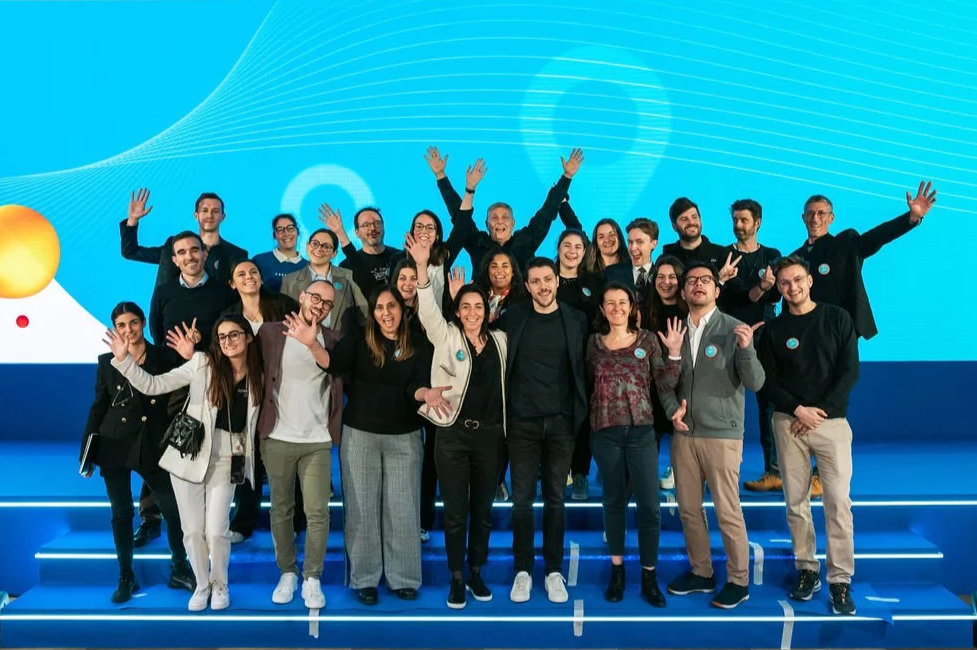 Group of diverse people gathered on stage, smiling and waving, with a blue digital-themed background.