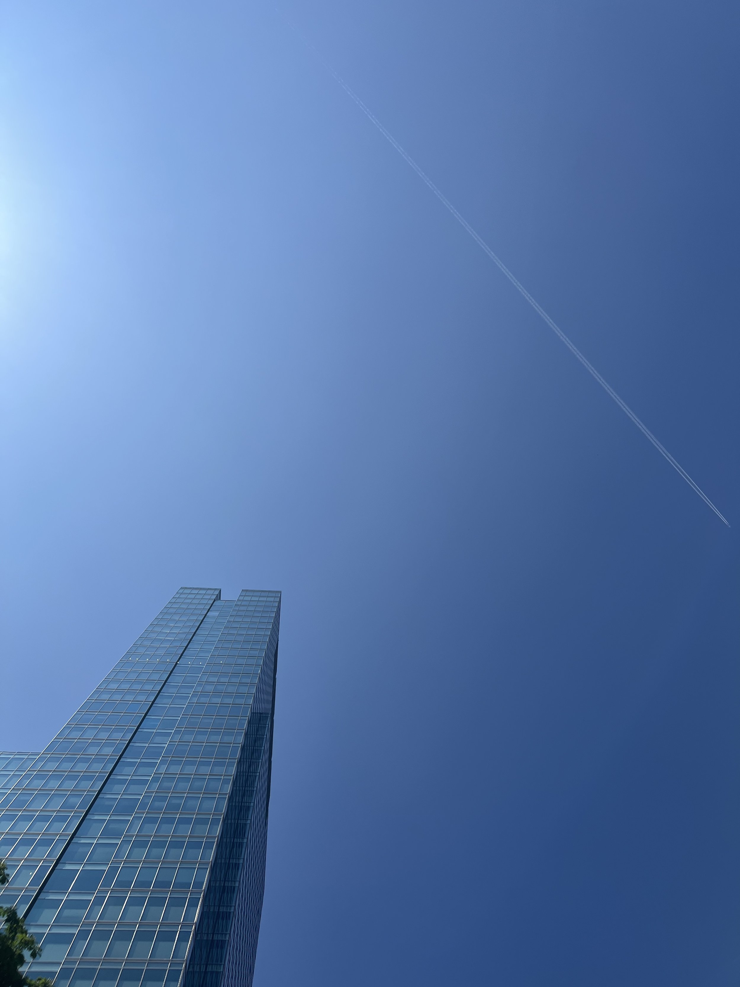 A tall, modern glass skyscraper with reflective windows against a clear blue sky, with a faint contrail from an airplane visible in the upper part of the sky.