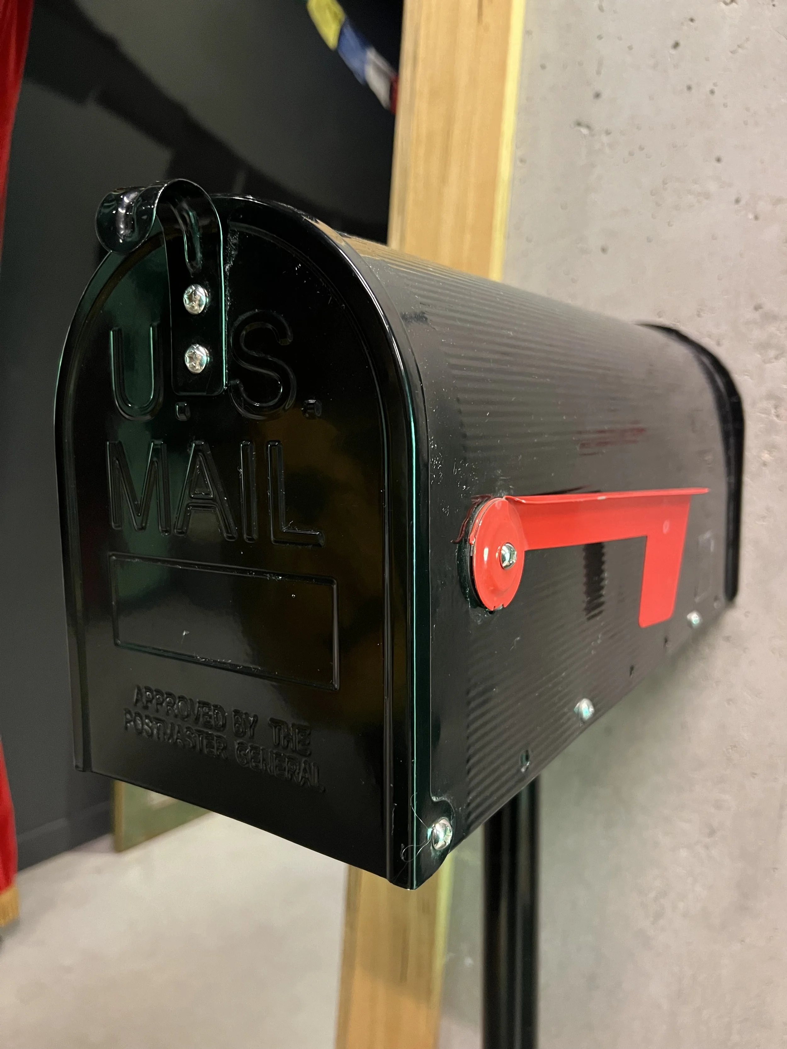 Close-up of a black mail collection box mounted on a wooden bench, with a red flag and metal screws visible.