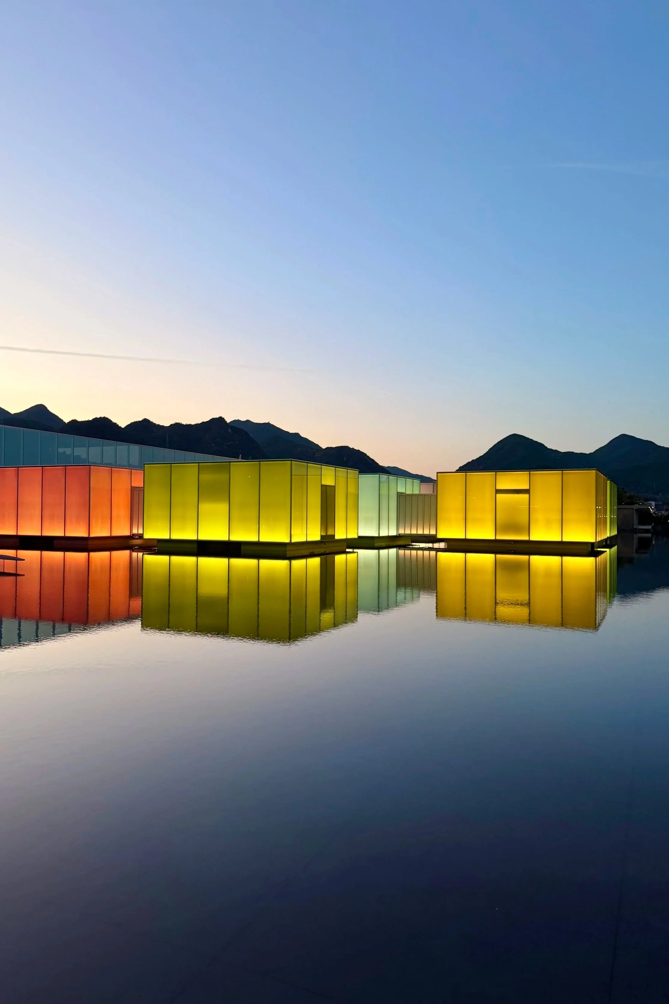 Colorful modern buildings with glass walls reflect on a calm body of water, mountains in the background, under a clear blue sky.