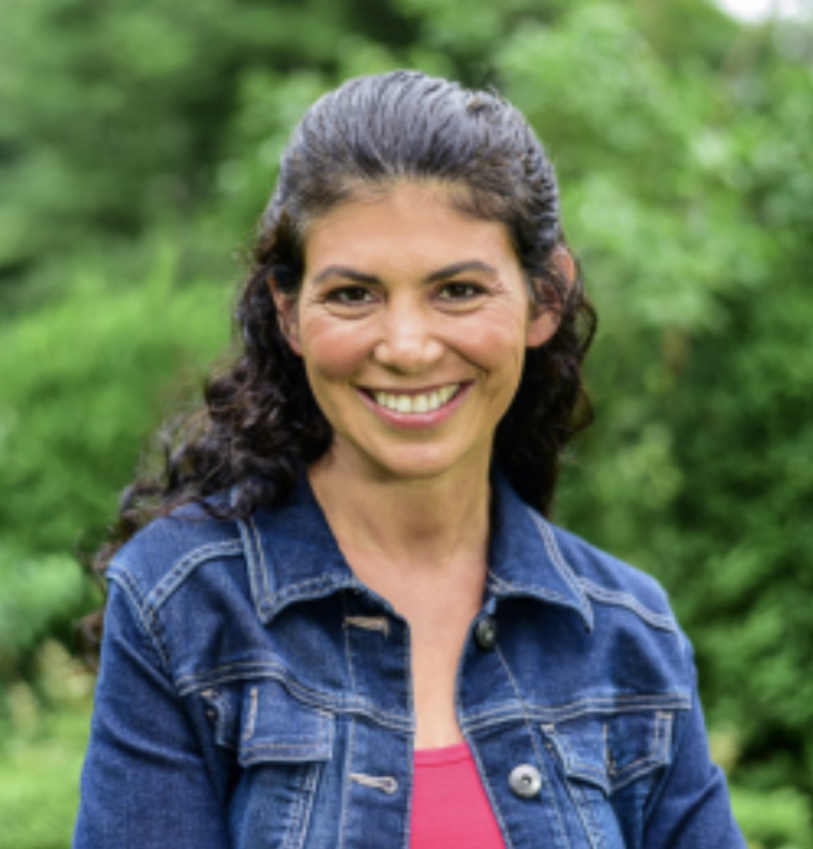 Teacher, Melissa Toole, woman with long, dark, curly hair smiling outdoors, wearing a denim jacket and a pink top, with a blurred green foliage background