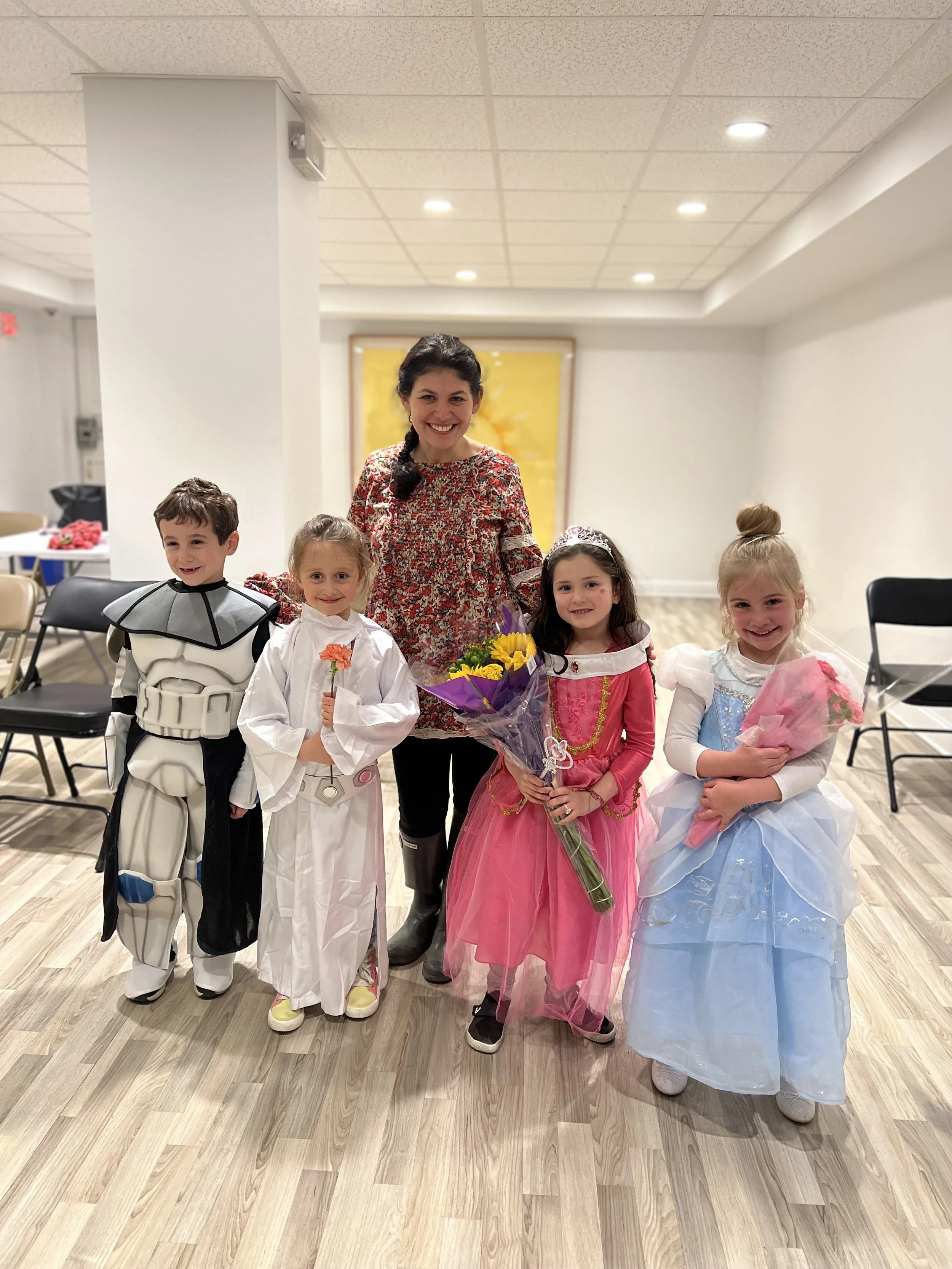 A woman and four children dressed in costumes, standing in a room with a wooden floor and white walls, smiling at the camera. The children are wearing costumes of a knight, princesses, and a fairy tale character, holding flowers, and the woman is standing behind them.