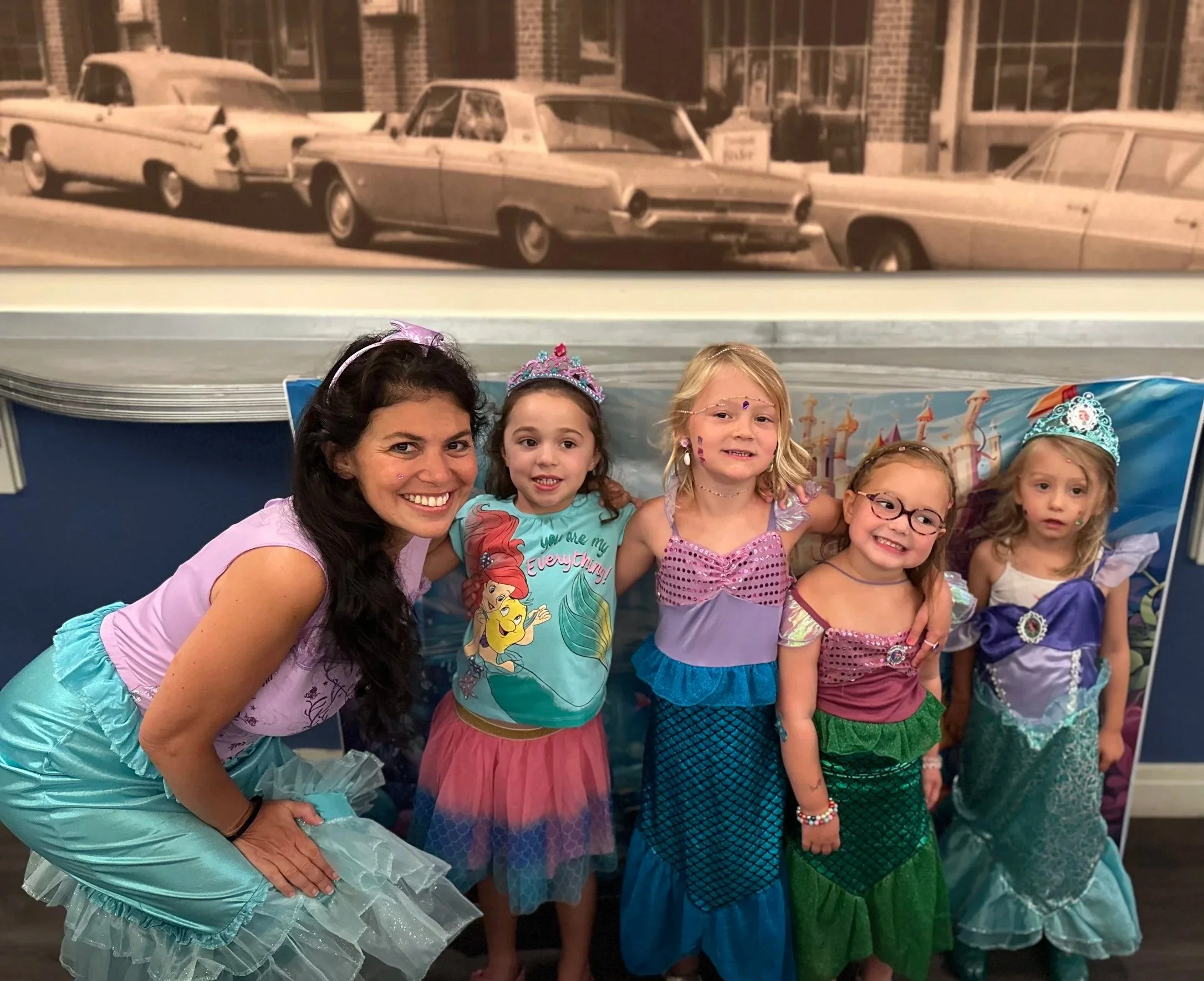 A group of children and an adult woman dressed as princesses or fairy tale characters at a party or costume event, standing in front of a colorful backdrop with a castle motif.